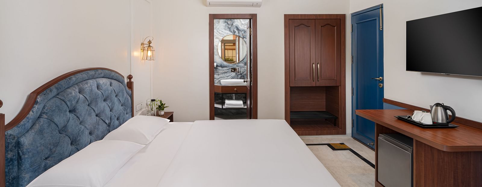 A side view of the room features a spacious bed, a wall-mounted television directly opposite the bed, and wooden furniture | Hotel Ganga Maiya