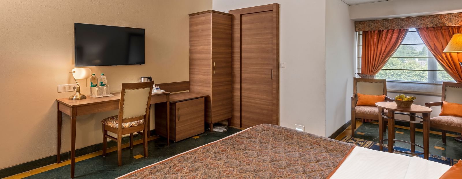 side view of a bedroom with a bed facing a television, below which is a workdesk and chair and a wardrobe beside it - The Manor, Kashipur