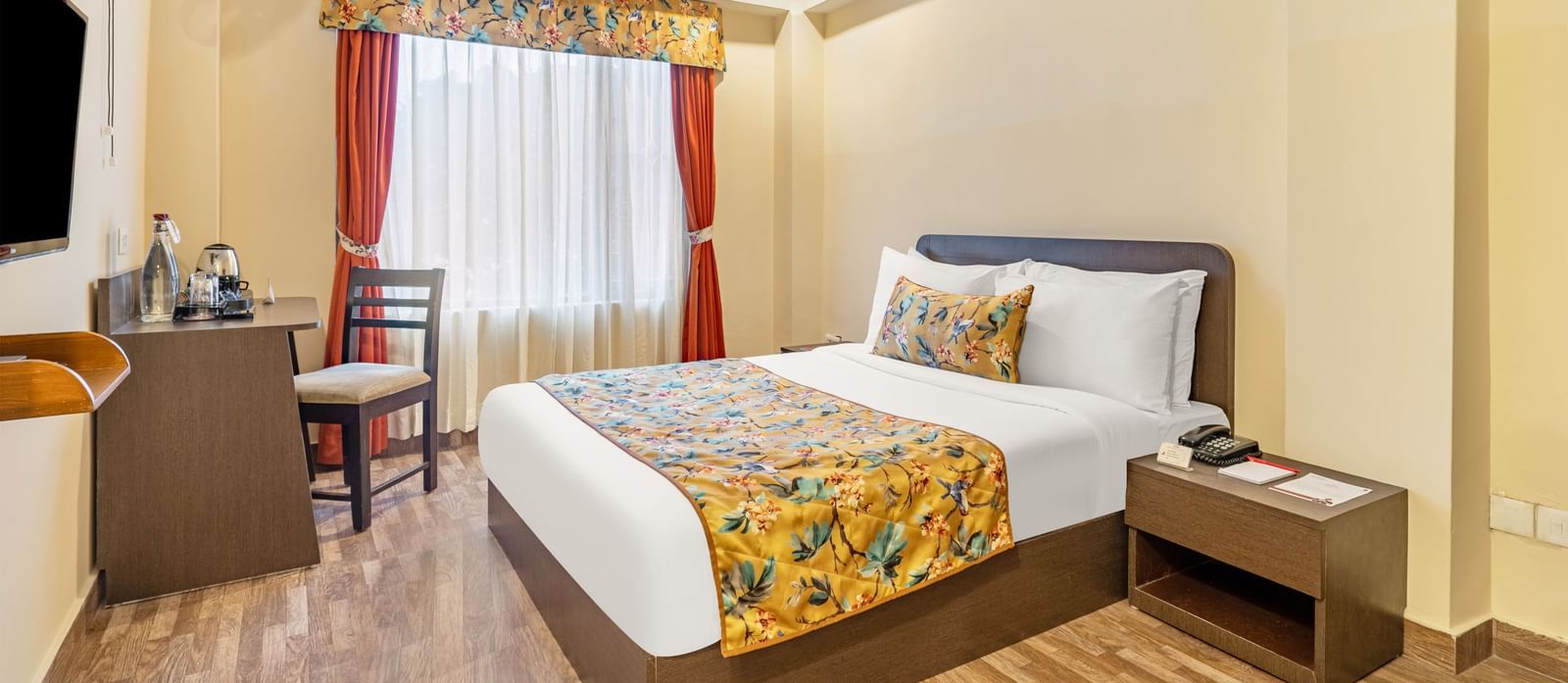 Well-lit hotel room with a double bed featuring floral bedding, a window with red curtains, a small work desk, wall-mounted TV, and wooden flooring - Summit Namnang Courtyard & Spa, Gangtok
