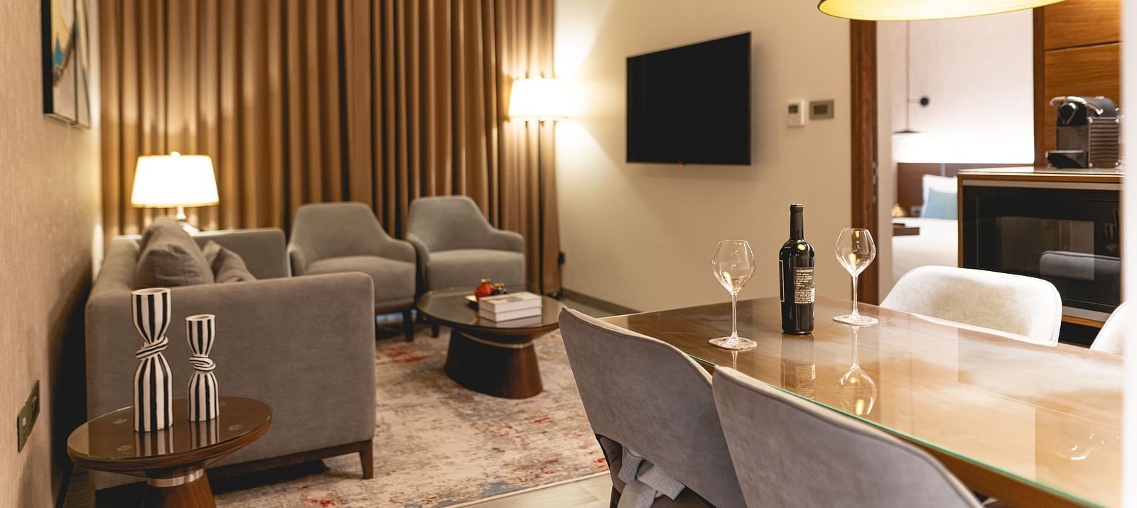 Modern living room at KertelSuites with neutral tones, featuring a sofa, armchairs, TV on the wall, floor and table lamps, and a dining table with chairs; warm lighting and vertical wooden slats on one wall