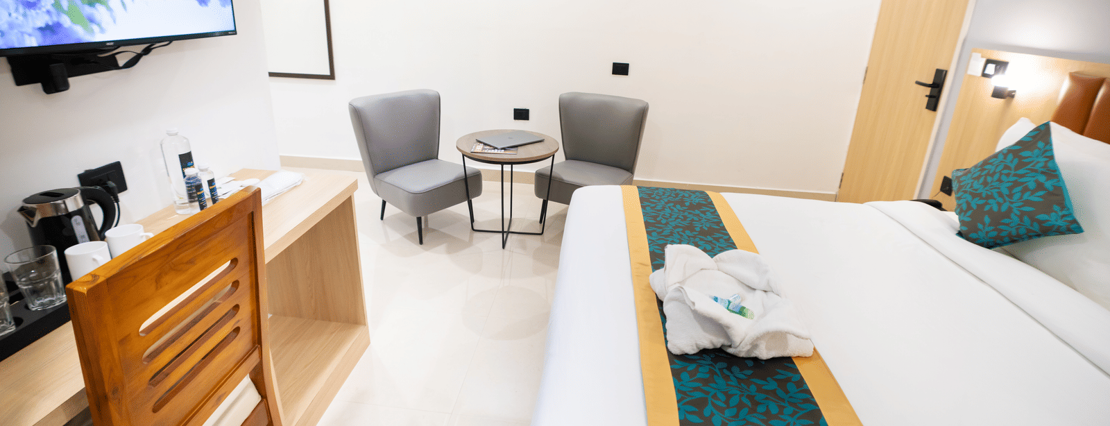 A view of the room with a large TV, a small table with chairs to the side, wooden interiors, and a spacious bed at Sunrise Business Hotel, Hyderabad.