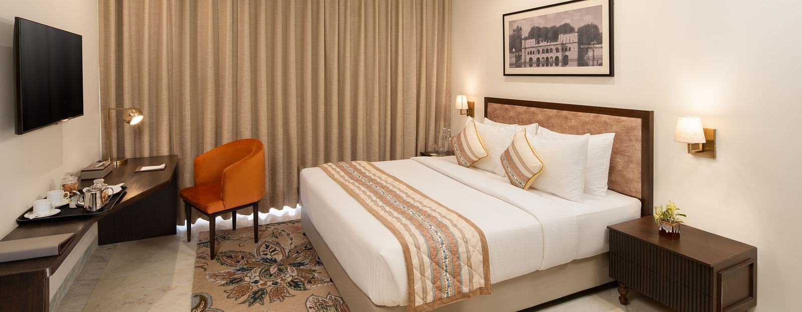 An image of a bedroom with bedside table, photo frame hanged on the wall, work desk with sofa chair, and a flat TV screen - Brij Eternity, Vrindavan