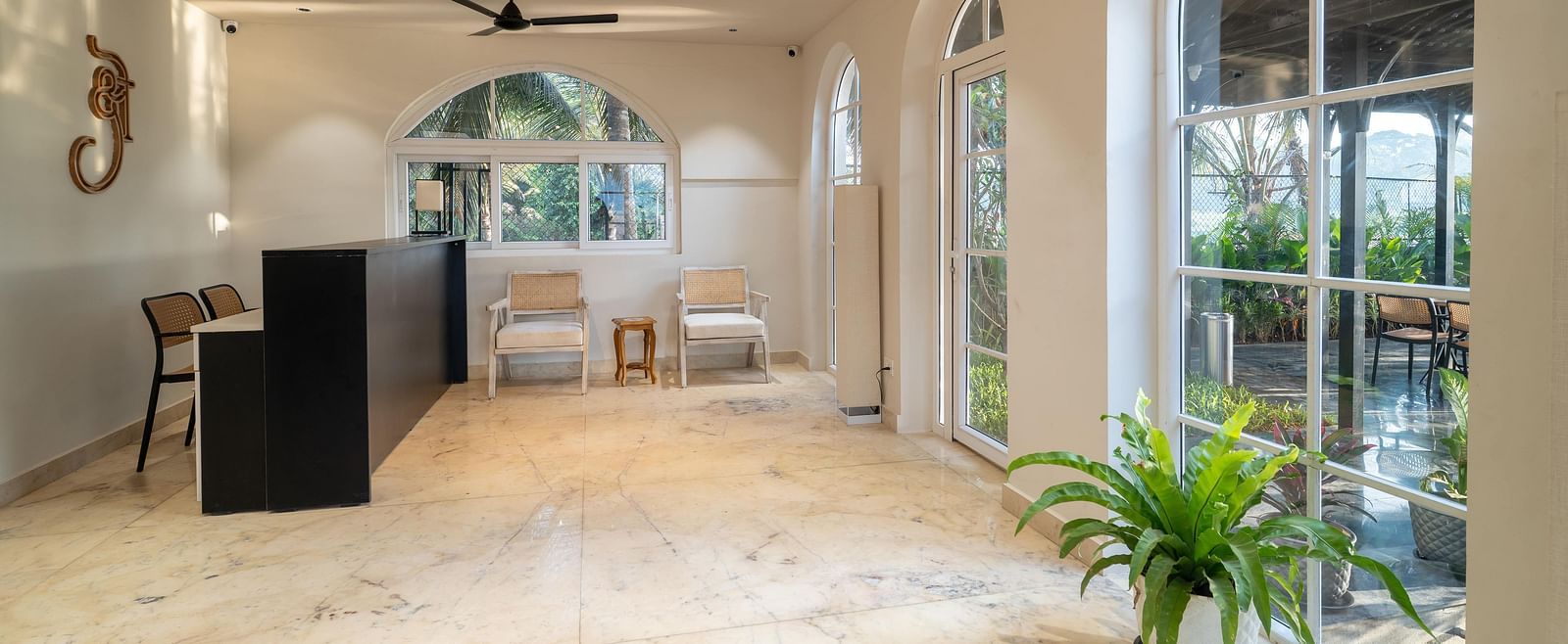 A bright reception space with a polished floor, leading towards arched windows showing greenery outside. A black front desk sits to the left, and a potted plant at Kssemya
