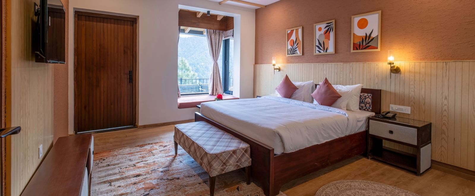 Spacious room with a large bed, bedside table, wall-mounted light above the headboard, a TV with a desk below, and windows and a door to the side at The Chamomile Estate.