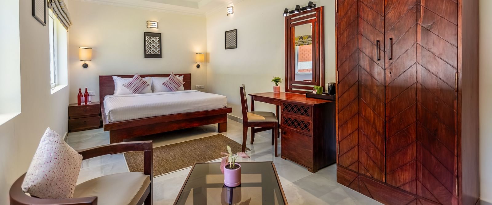 A bright, clean hotel room with a bed, a wooden desk and chair, a large wooden wardrobe, and a seating area with a glass-topped coffee table - Mango Hill Resort, Coimbatore
