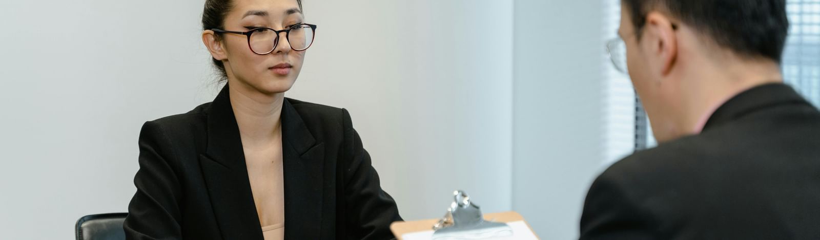 Two professionals with black suits are engaged in a  interview at an office desk with a clipboard. 