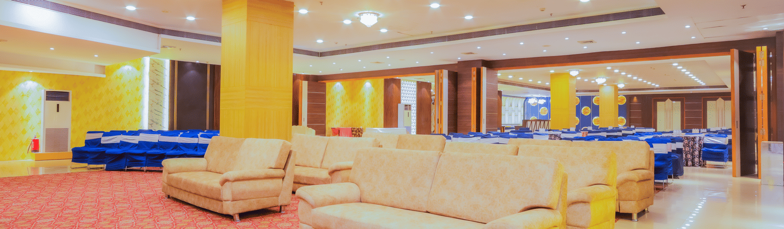 A large, open hotel lobby or lounge area featuring cream-colored sofas and chairs on a red patterned carpet, with bright lighting and decorative elements.