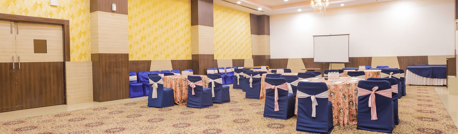 A spacious conference or event room with a patterned carpet, featuring chairs draped in blue covers arranged around tables, and a stage area.