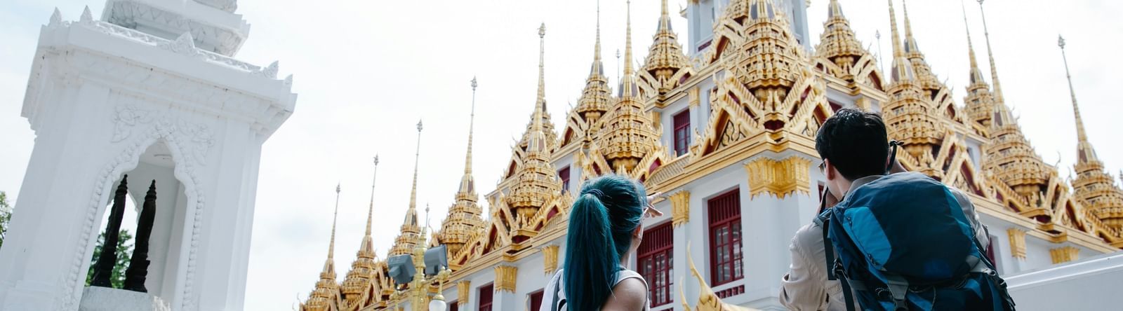 Two tourists with backpacks view a golden-roofed temple in Thailand, with an ornate white structure in the foreground.