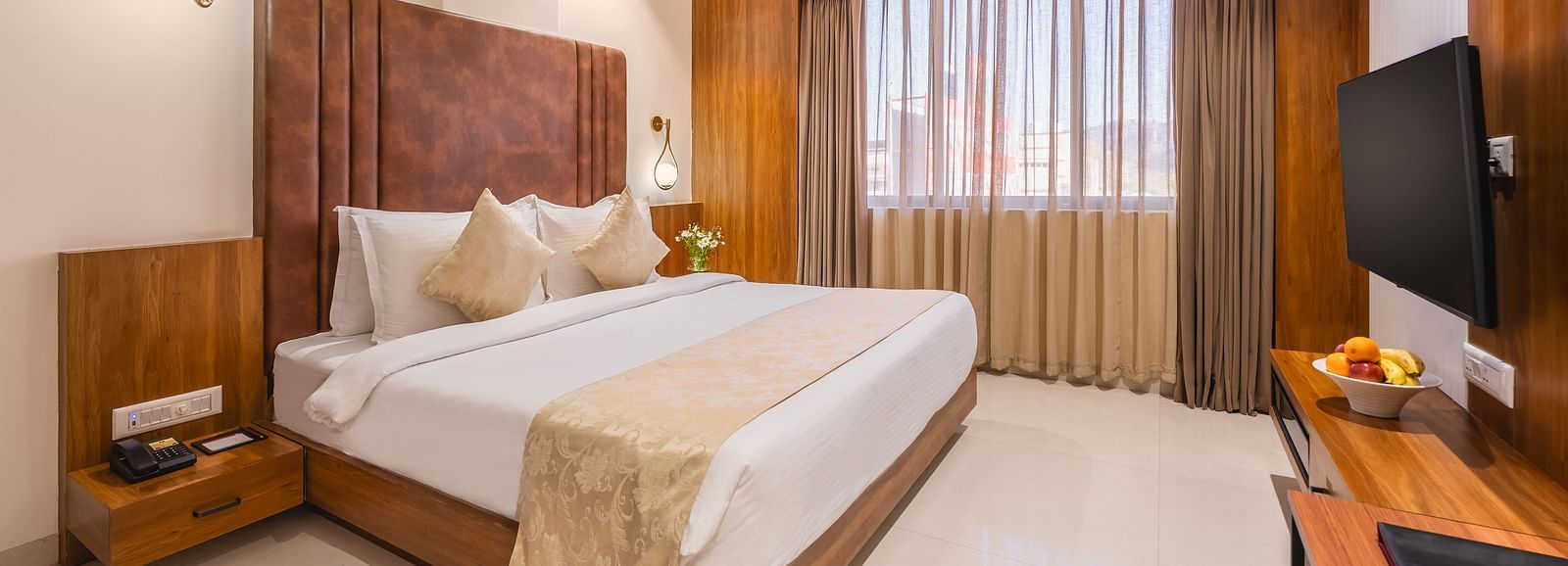 A comfortable king-size bed with soft pillows and a decorative throw faces a mounted television in a well-lit hotel room with a window - Lords Eco Inn Ambaji.