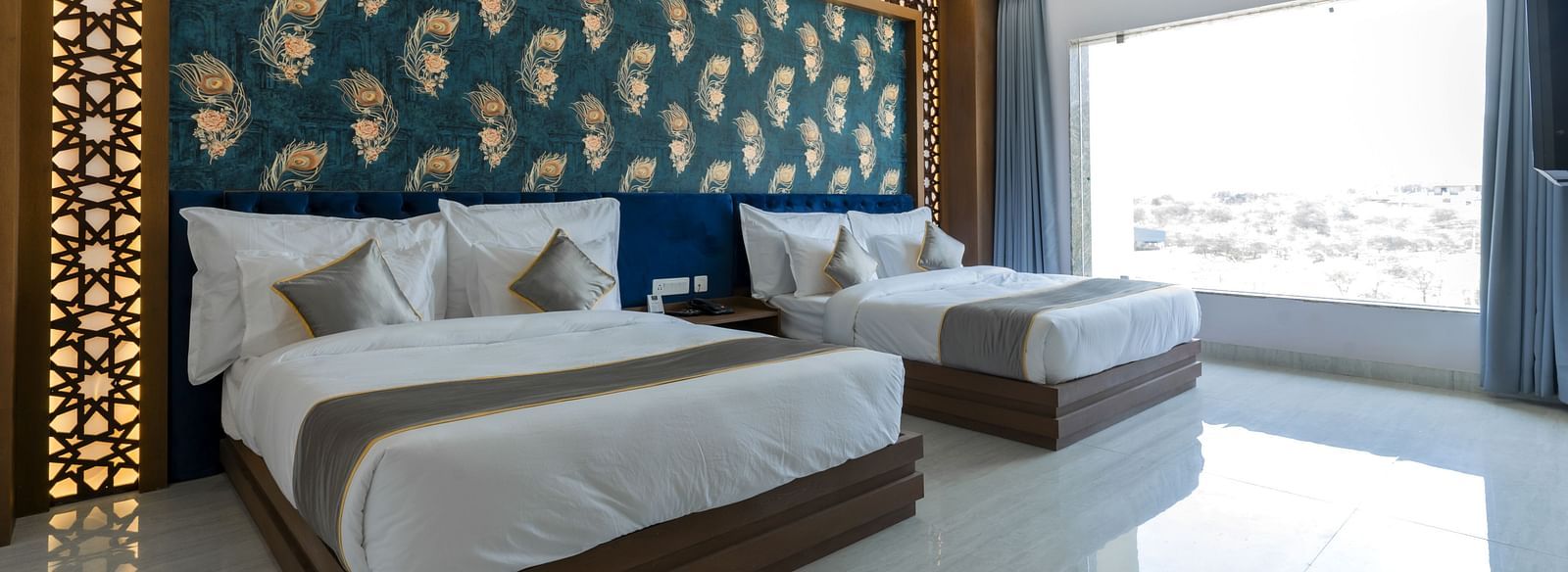 A bedroom at the Bamboo Saa Mulberry Resort featuring two twin beds with white linens and grey throws. A decorative wall panel with a blue and gold pattern is above the beds and a large windows is visible.