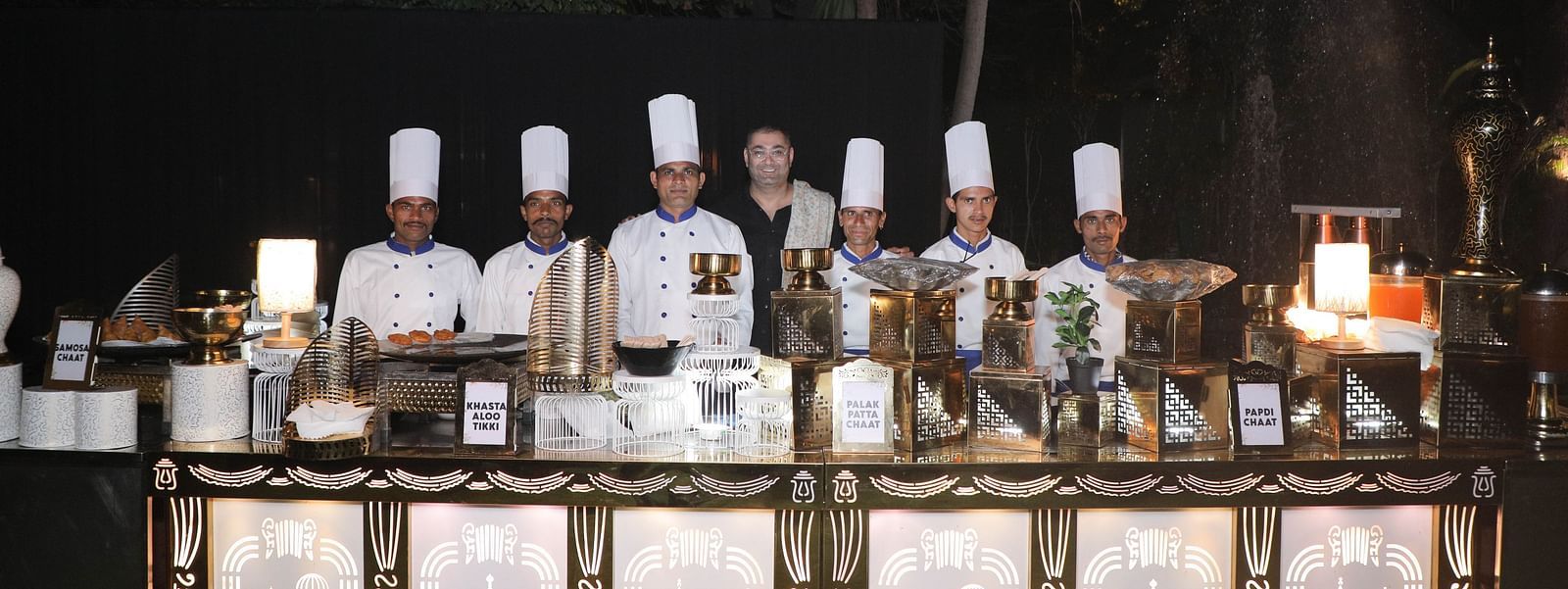 Chefs presenting dishes at the buffet counter under the night sky, with open-top dining set up at the 'Around The World in Eighty Plates' event.