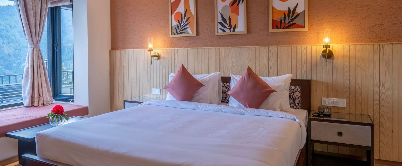 A view from the foot of the bed at The Chamomile Estate, showcasing bedside tables with wall-mounted lamps on either side of the headboard, and a window with curtains drawn aside.