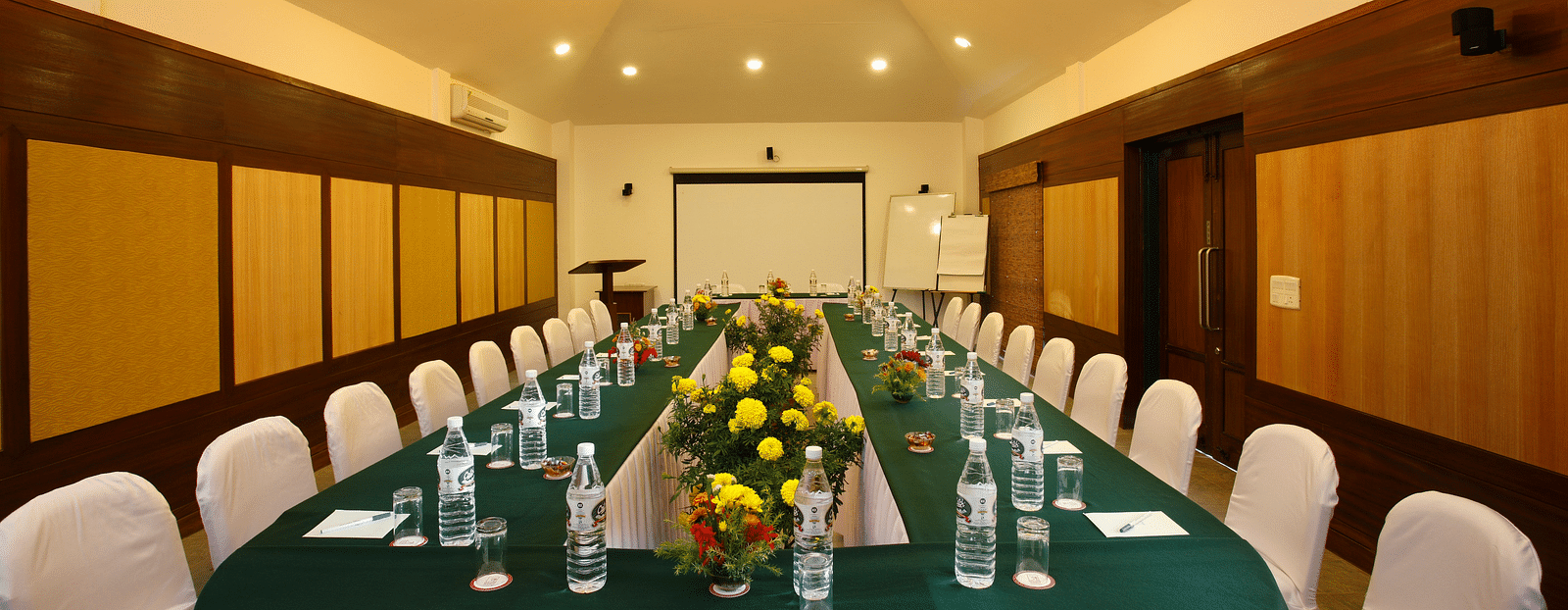 A spacious meeting hall featuring a large table set with water bottles and floral centre pieces, surrounded by neatly arranged chairs -  Baikunth resort, kasauli 