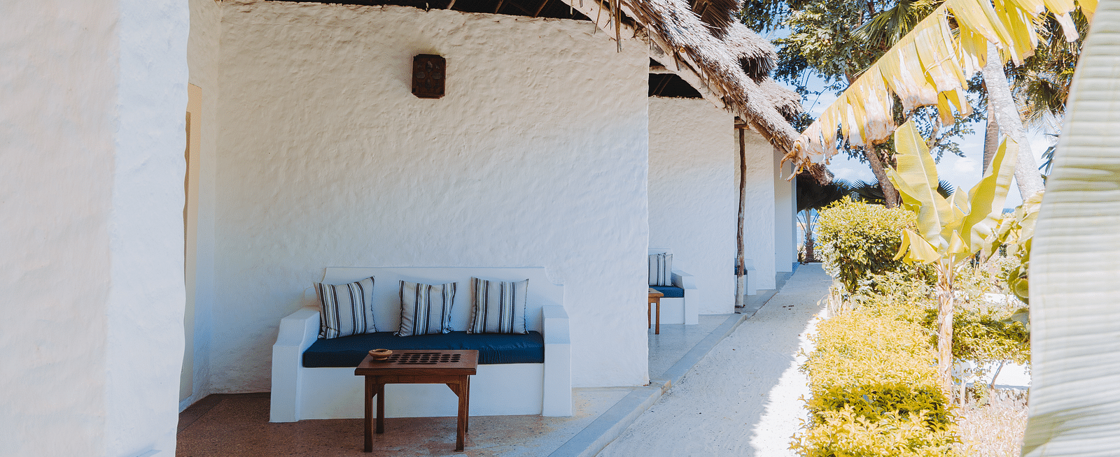 Exterior view of a building with a thatched roof overhang, featuring a small patio with a cushioned bench and a wooden table - Shooting Star Boutique Hotel.