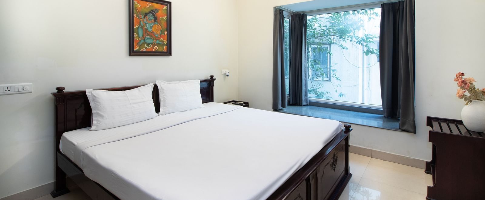A room at Sanctum Suites Indiranagar, Bangalore, offering a double bed with white linens, a bedside table with a lamp, a window letting in natural light, and a framed picture on the wall.