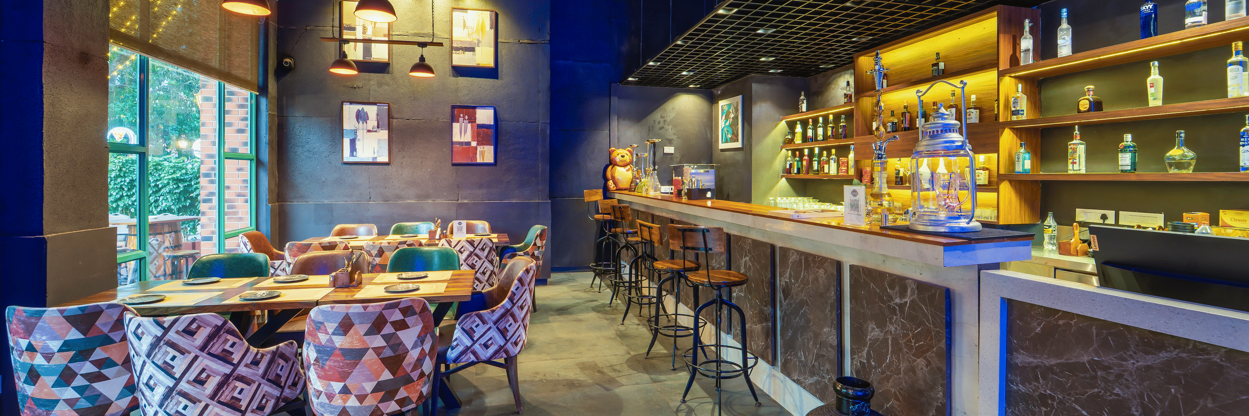 Rosetum Anjuna's Speakeasy Bar showing bar counter with wooden stools, patterned upholstered chairs, backlit bottle shelves, framed wall art, and blue-toned ambience.