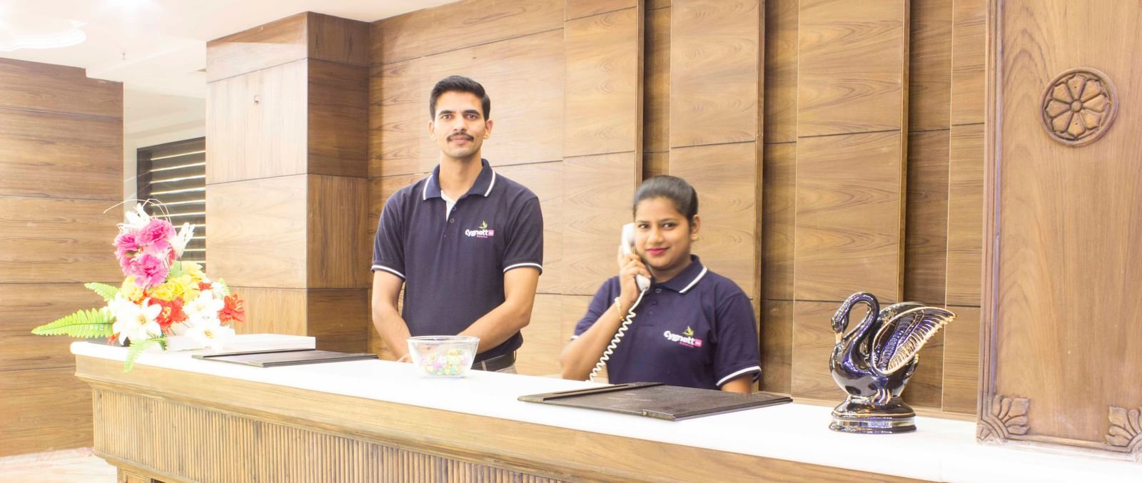 A hotel reception desk with two staff members smiling | Daksh The Madhuvan Suites