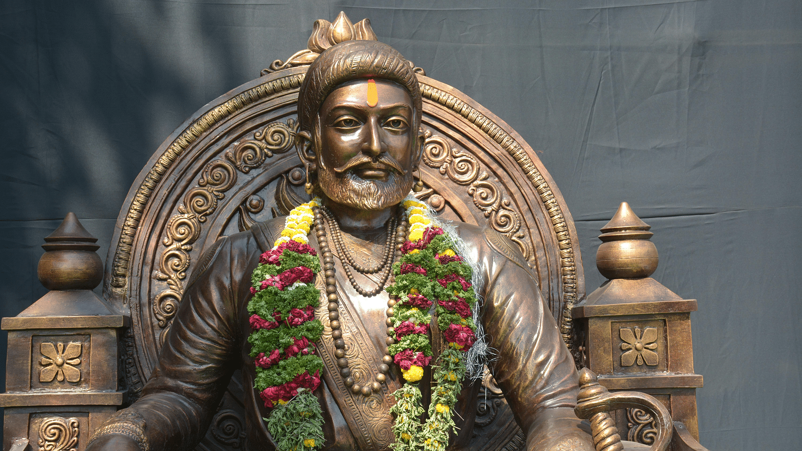 Bronze statue of Chattrapati Shivaji Maharaj sitting calmingly magnificently, decorated with garlands and flower decors.