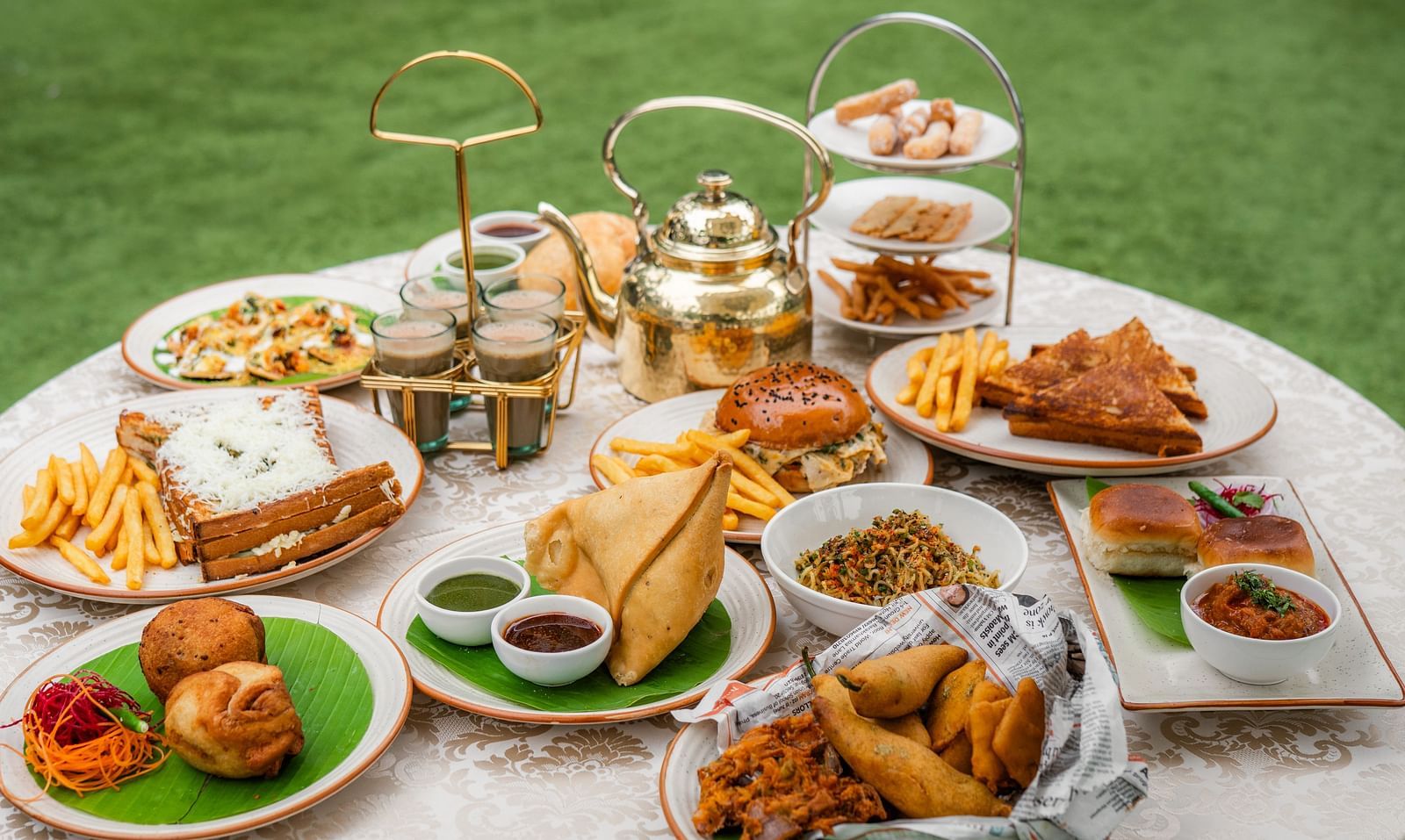 A breakfast table where varieties of savouries served in each plat consisting of samosa, french fries, sandwiches, salsa, fritters, bun, etc. There is also a kettle and hot tea served in four cups. 