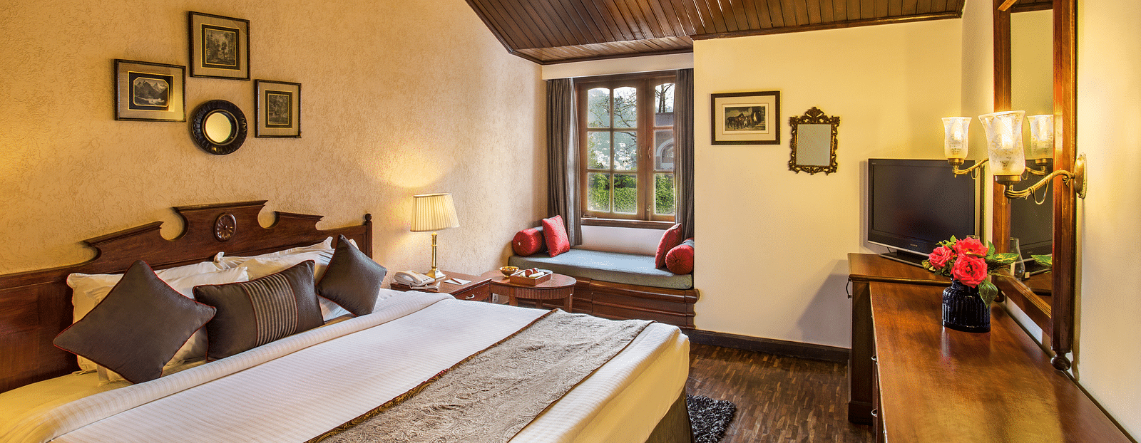 An image of a cozy room in naini retreat, nainital, with a king-size bed, wooden furniture, and a bay window overlooking a garden. The room features a ceiling fan, a television, and a small sitting area.