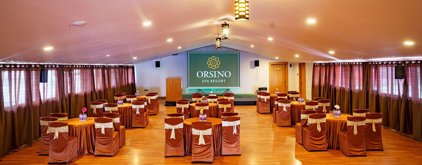 A spacious event hall at Orsino Spa Resort with tables draped in gold fabric and chairs, under a high ceiling with multiple hanging lights.