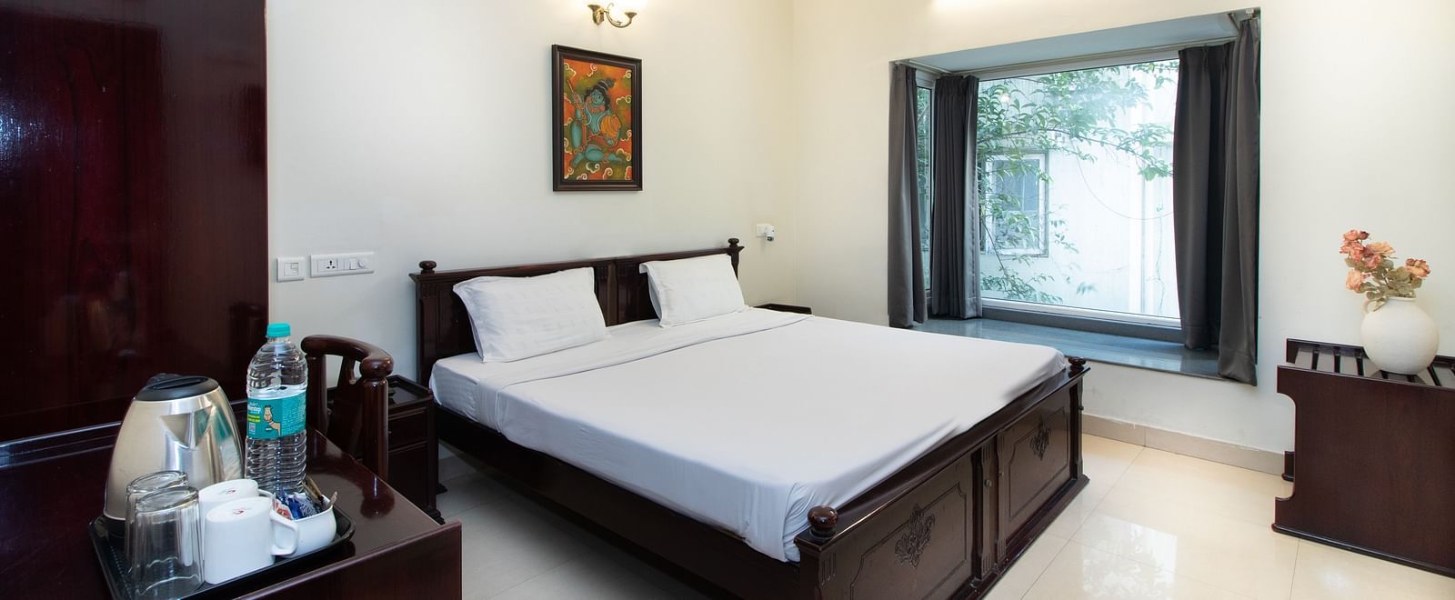 A room in Sanctum Suites Indiranagar, Bangalore, showcasing a double bed with white bedding, a bedside table with a lamp, an electric kettle placed on a table, a chair, and a window providing natural light.