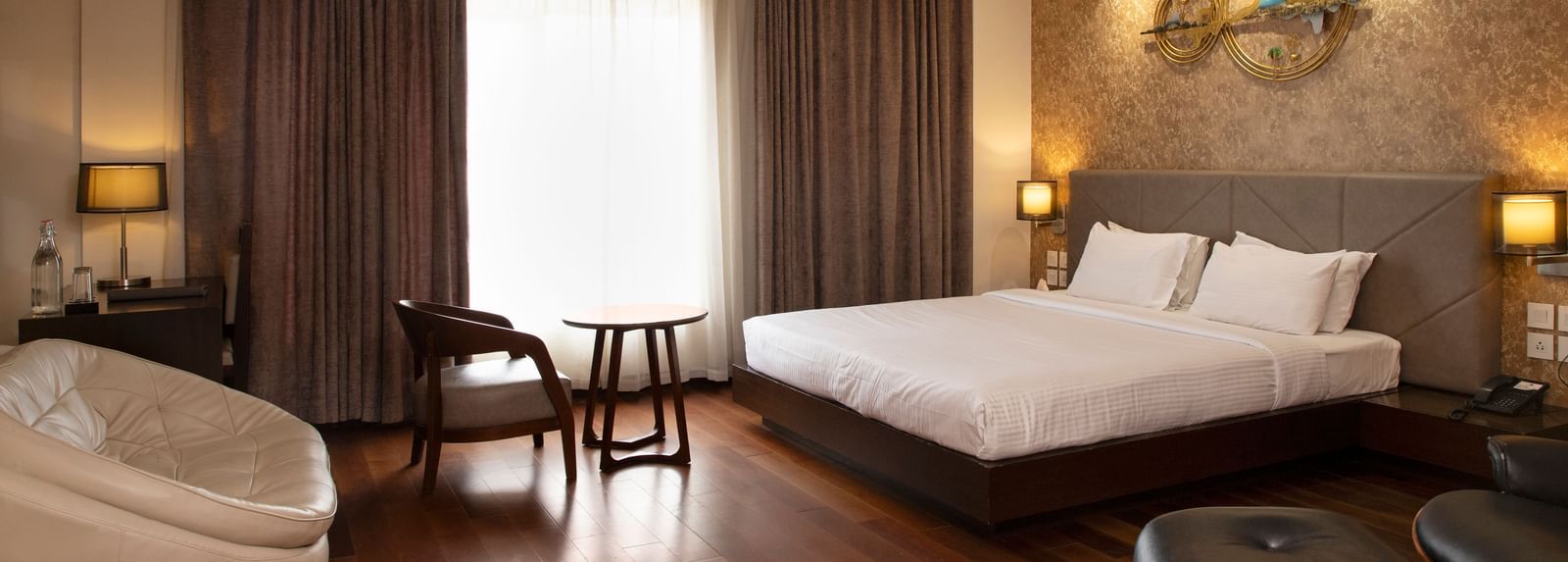 A spacious room featuring a large bed, a cosy seating area, elegant curtains, soft ambient lighting, and expansive windows with curtains drawn to the side at The Hamuse Hotel, Kodaikanal.