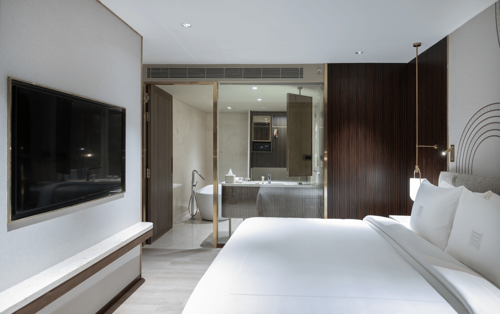 Bedroom with a plush bed facing a TV on the wall and a glass partition to the bathroom area at Merlis Hotel, Coimbatore