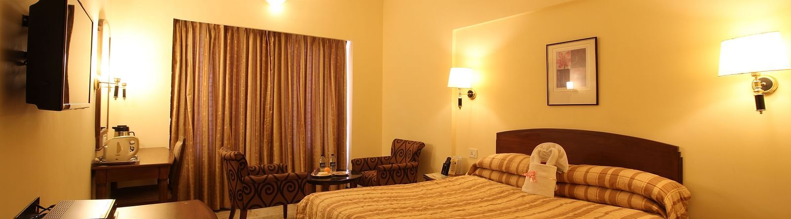 A room with a large bedding with wall lamps on either side of the bed, coffee table and chairs, on a bright lighted setting.