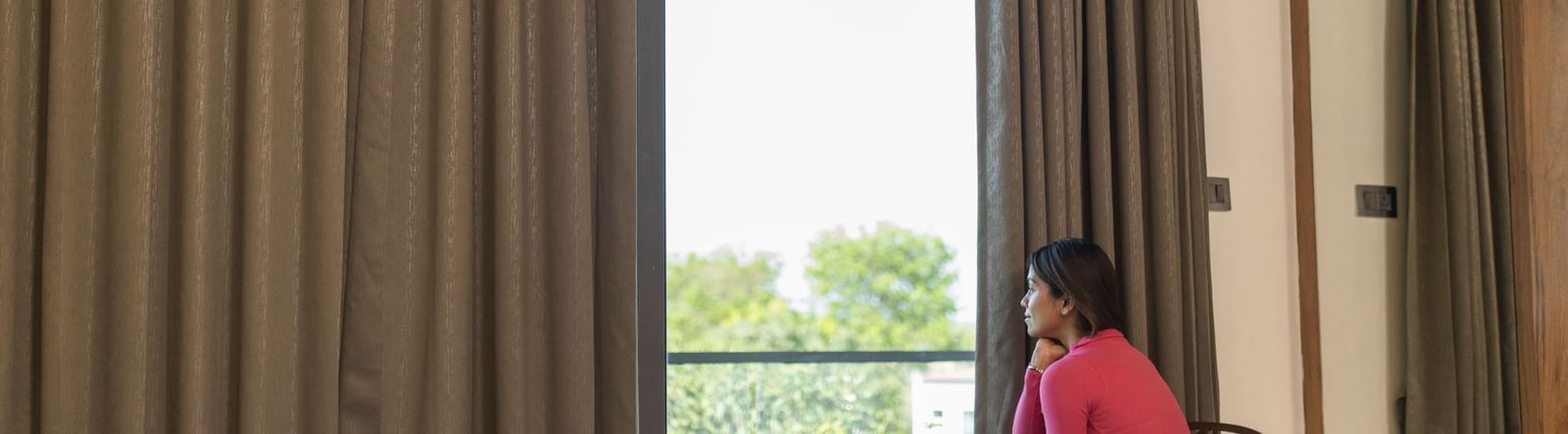 A girl sitting on a double bed looking at the view outside of the Daksh Comfort Room at Daksh Eden Greenz, Sasan Gir.