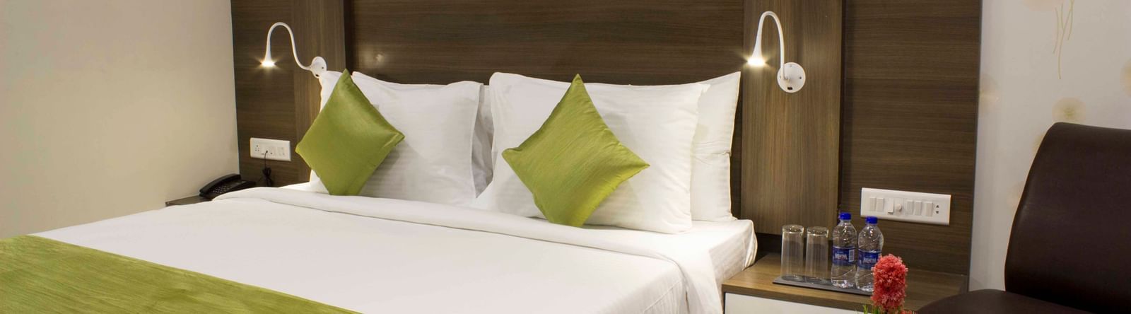A hotel bedroom with a neatly made bed, green accents, and bedside tables with lamps | Daksh Comfort Room | Daksh The Madhuvan Suites