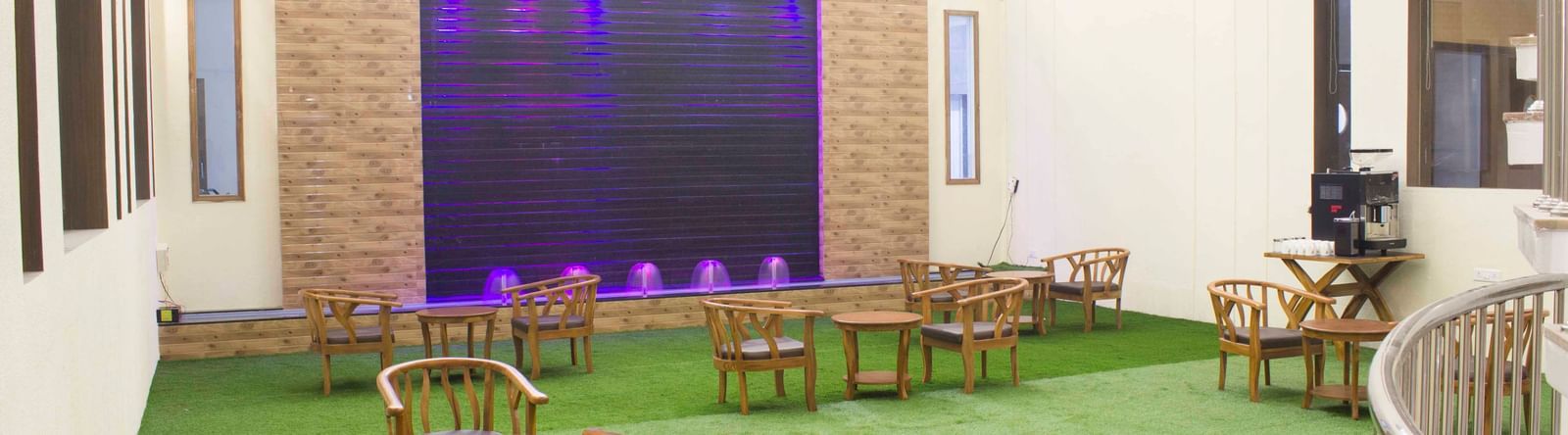 An indoor area with wooden chairs and tables on green artificial grass, facing a water wall | Daksh The Madhuvan Suites