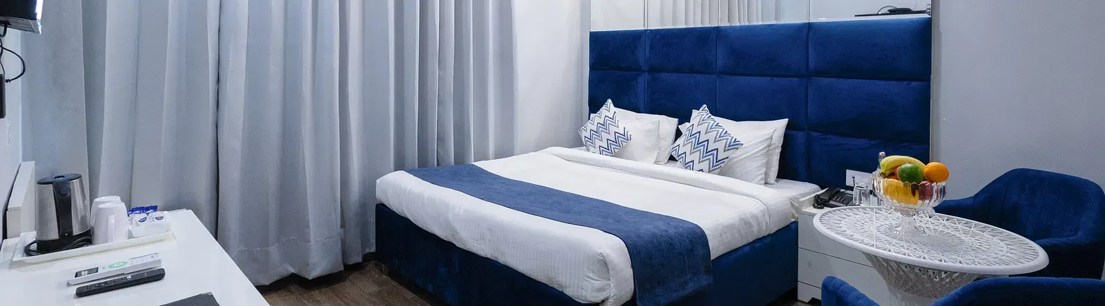 Deluxe Room featuring a king size bed with a blue headboard and white and blue linens, two blue chairs with a small round table, and light wood-look flooring. - Hotel Downtown17, Chandigarh.