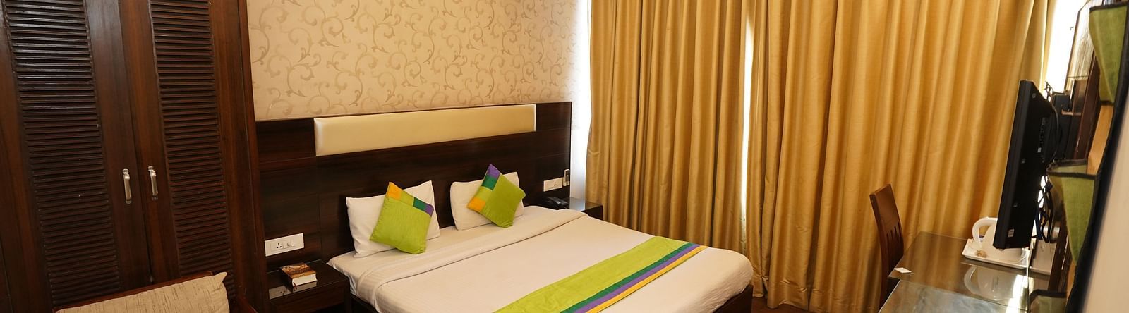 Deluxe Room featuring a king size bed with green accents, a wooden headboard, bedside tables, curtains, a desk, and a chair.- Hotel Dreamland, Chandigarh