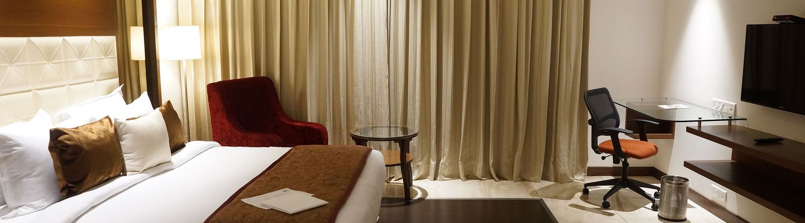 Shot of a king size bed in a spacious standard room featuring a flat screen television, a desk and a chair and bed side lamps  | Marigold Hotel Jaipur 