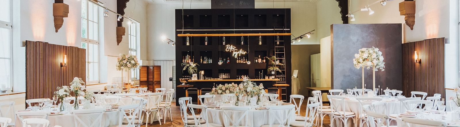 Elegantly set dining hall with white chairs, round tables with white linens and a central bar area under rustic industrial beams