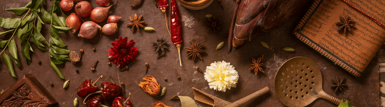 Assorted spices and kitchen tools beautifully arranged with flowers and vegetables on a rustic background | Kamats India