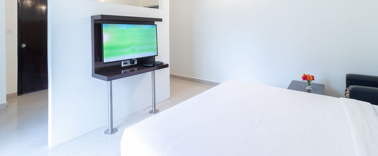 A room with a double bed, a wall-mounted television, and a glimpse into the other part of the room at Sanctum Suites Domlur, Bangalore.