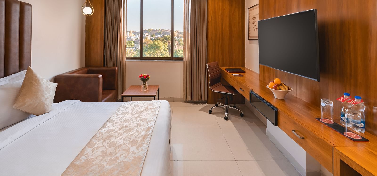 A well-furnished hotel room includes a comfortable bed, a seating area, a desk with a chair, and a mounted television - Lords Eco Inn Ambaji.
