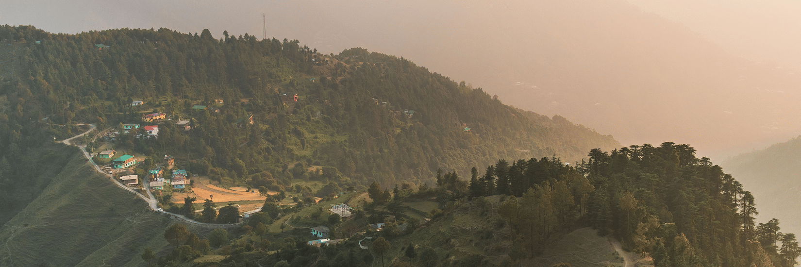 Panoramic view of forested hills in Kasauli, winding mountain roads, and scattered buildings across a hillside, with layered mountain ranges in the distance.