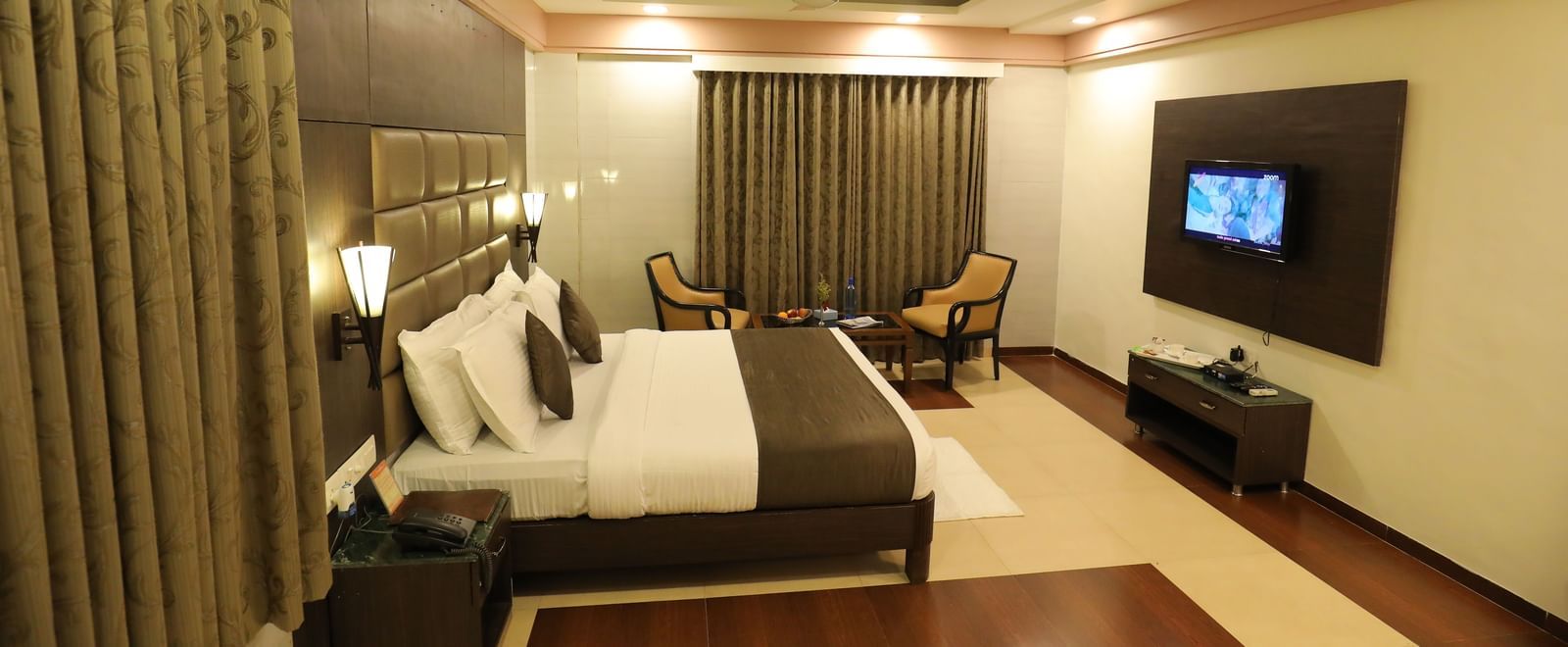 A wide view of a bedroom at Krushnai Resort with a large bed, lounge chairs and large windows with drapes closed.