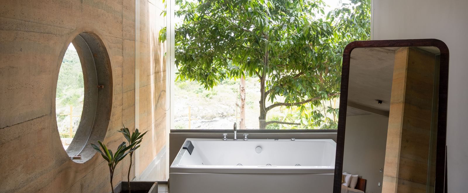 A bathroom featuring a large round mirror, a vanity area, and a window overlooking lush trees | Gaia Studio | RD's Nature Retreat, Bangalore