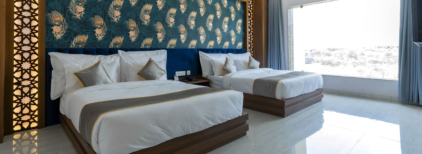 A bedroom at the Bamboo Saa Mulberry Resort featuring two twin beds with white linens and grey throws. A decorative wall panel with a blue and gold pattern is above the beds and a large windows is visible.