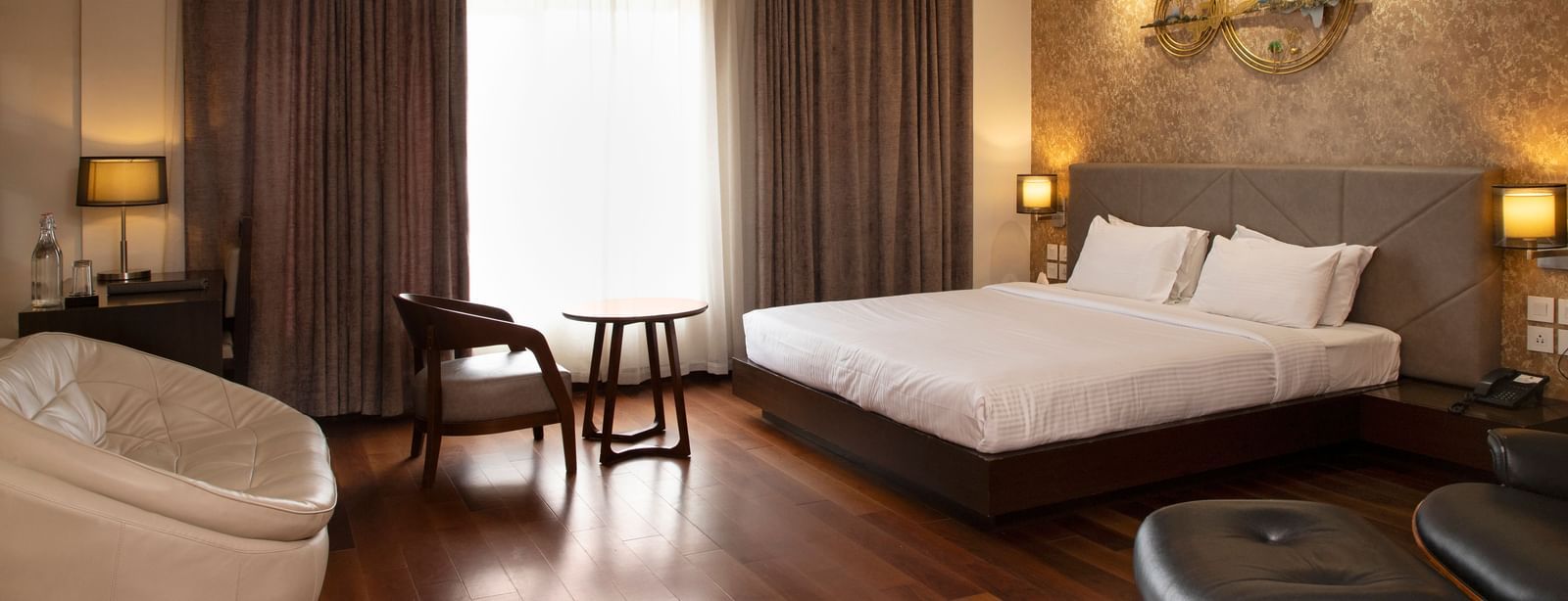 A spacious room featuring a large bed, a cosy seating area, elegant curtains, soft ambient lighting, and expansive windows with curtains drawn to the side at The Hamuse Hotel, Kodaikanal.