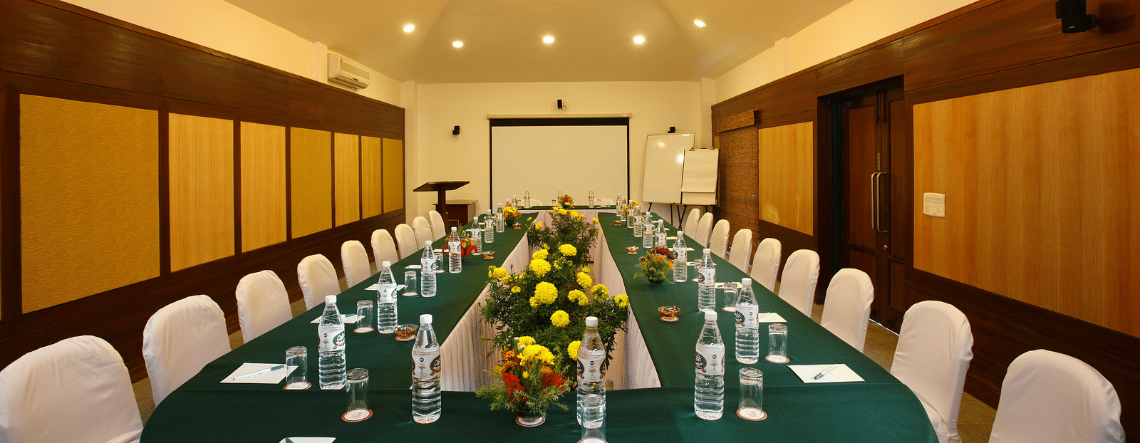  A spacious meeting hall featuring a large table set with water bottles and floral centre pieces, surrounded by neatly arranged chairs -  Baikunth resort, kasauli 