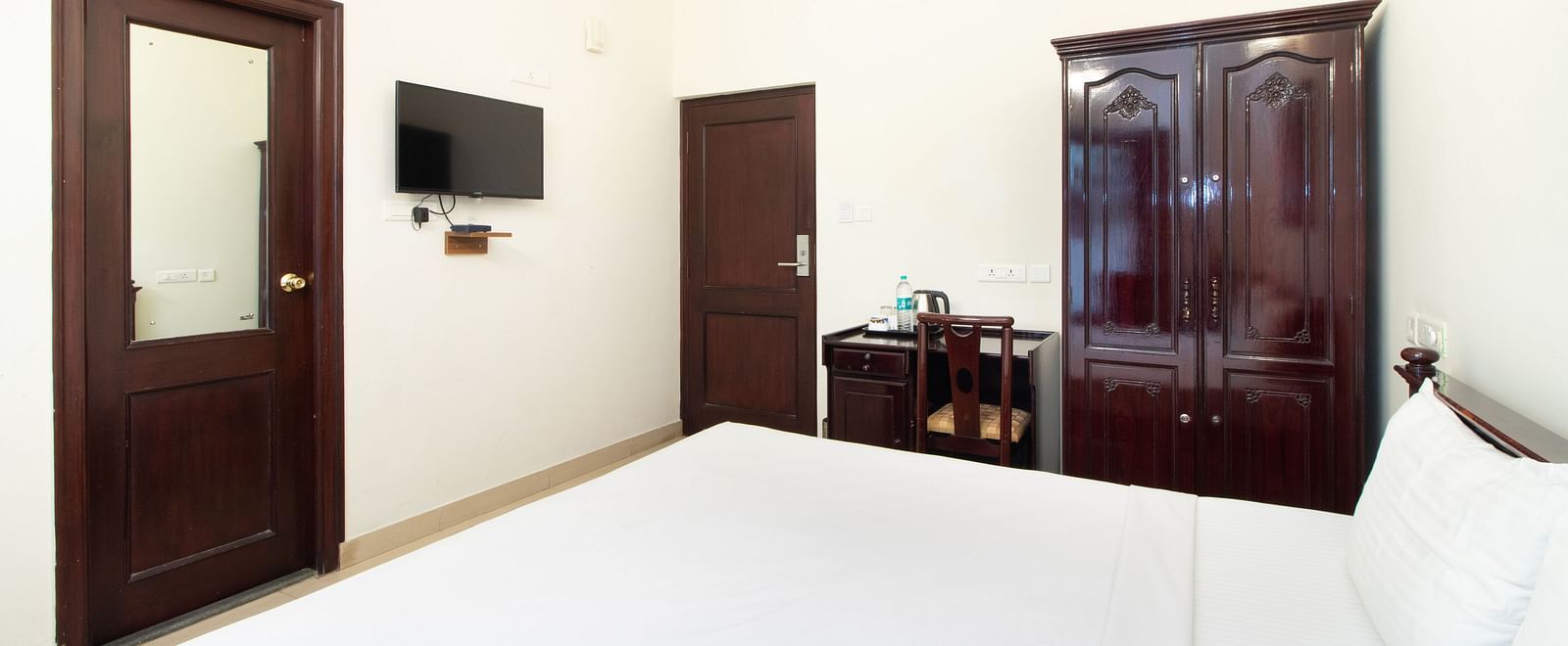 A bright room at Sanctum Suites Indiranagar, Bangalore, featuring a neatly made double bed, wooden furniture, and a wall-mounted television.