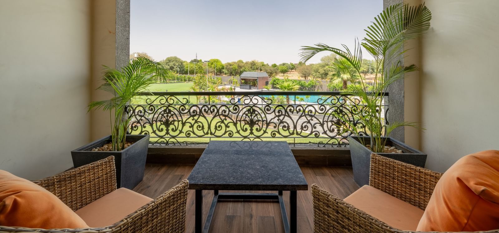 A balcony of executive king room in Hotel Highway King, Bagru, featuring cushioned lounge chairs and a coffee table, overlooking lush garden under an overcast sky.