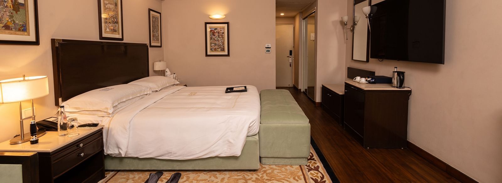 image of a brightly lit room with a king size bed and a wall mounted television placed opposite to the bed at our heritage balcony room - image of the heritage balcony room with sleek interiors and a a plush king size bed - The Claridges New Delhi
