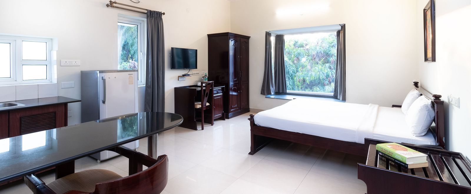 A spacious room offering a double bed with white linens, a dining table with chairs, a television, a refrigerator, and a window providing natural light at Sanctum Suites Indiranagar, Bangalore.